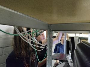 Drie ULYSSIS-leden staan achter een rek met kabels, monitors en servers. Ze zijn aan de kabels aan het werken.